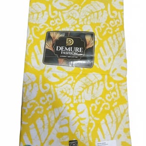 May include: A folded piece of bright yellow fabric featuring a white leaf pattern. A black label in the center reads DEMURE FASHION 6 YARDS 100% COTTON. A small circular sticker in the corner displays text and the words DE DEIER FES E COLORS.