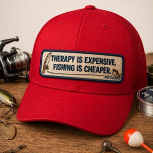 May include: A red baseball cap with a mesh back and a rectangular patch. The patch reads "THERAPY IS EXPENSIVE. FISHING IS CHEAPER." in white text with a fishing rod and fish graphic. Fishing gear is visible in the background.