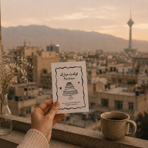 Puede incluir: Una tarjeta de felicitación con una ilustración de pastel y texto en farsi se sostiene frente a un paisaje urbano. Una taza de café se encuentra en una repisa junto a la tarjeta. Un jarrón con flores blancas también está en la repisa. El fondo presenta un horizonte y una torre.