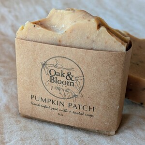 May include: A rectangular bar of tan-colored soap with visible dark specks, wrapped in a brown paper label. The label features the text Oak & Bloom Farm, PUMPKIN PATCH, Handcrafted goat milk + herbal soap, and 4oz, with a circular botanical logo.