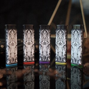 May include: Five rectangular lighters featuring a black body with a white intricate graphic print of a horned creature and a pentagram. Each lighter has a distinct coloured base in blue, red, purple, yellow, or green, with a black top ignition mechanism.