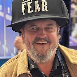 May include: Black baseball cap with the text "TRUTH OVER FEAR" in white block letters. The person in the image is wearing a tan jacket over a black and white patterned shirt.