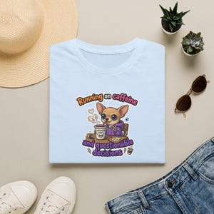 Op de afbeelding: Een lichtblauw t-shirt met een afbeelding van een chihuahua in een paarse hoodie die aan een bureau zit met een kop koffie. De tekst luidt Running on caffeine and questionable decisions. Het ontwerp bevat stoom, een klein kopje en bliksemschichten.