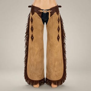 May include: Tan suede leather chaps featuring a dark brown leather waistband with a silver-toned metal buckle. The legs are adorned with dark brown diamond-shaped leather inlays and long, flowing fringe trim along the outer seams of each leg.