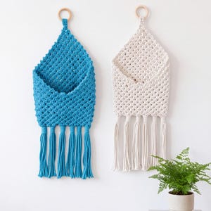 May include: Two wall-hanging macrame pockets, one in teal and one in off-white, each featuring a diamond-patterned weave, a wooden ring at the top for suspension, and five long, dangling tassels at the bottom edge for a textured finish.