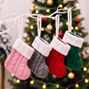 May include: Four small Christmas stockings hanging from a white branch. Each stocking features a cable knit pattern in pink, gray, red, or green, with a white ribbed cuff and a matching white fabric loop for hanging on a tree or display.