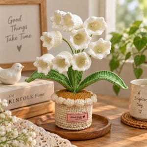 Op de afbeelding: Een gehaakte lelietje-van-dalen plant met witte klokvormige bloemen en groene bladeren, in een beige gehaakte pot met een geschulpte rand en een roze rechthoekig label met de tekst Hand Made with love.