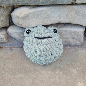 May include: A round, sage green crochet plush toy shaped like a frog. The stuffed creature features a textured, chunky yarn surface, two small black plastic eyes positioned near the top, and a single horizontal black stitched line for a mouth.