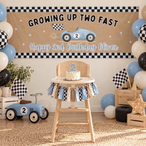 May include: A rectangular banner with a tan background, black and white checkered borders, and the text "GROWING UP TWO FAST" above a blue race car illustration and "Happy 2nd Birthday Oliver" in blue script, displayed with balloons.