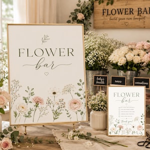 Può includere: Un cartello rettangolare con cornice dorata che presenta il testo "FLOWER bar" in nero, decorato con illustrazioni ad acquerello di fiori rosa e bianchi e fogliame verde. Un cartello più piccolo abbinato si trova nelle vicinanze con testo.