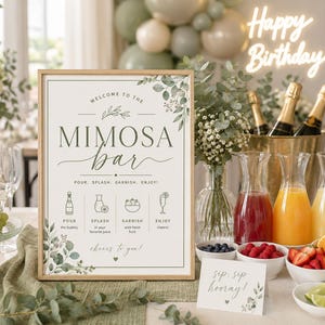 May include: A rectangular sign in a light wood frame with the text "WELCOME TO THE MIMOSA BAR" and "POUR. SPLASH. GARNISH. ENJOY!" featuring green eucalyptus illustrations, four instructional icons, and the handwritten-style text "cheers to you!" at the bottom.