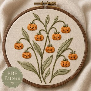 May include: An embroidery hoop containing a cream-colored fabric with a stitched design of green stems and leaves featuring eight small orange pumpkins with embroidered smiling faces. A circular green label reads PDF Pattern.
