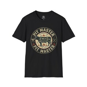 May include: A black short-sleeve t-shirt featuring a circular graphic on the chest. The design includes a cow diagram with meat cuts, flames, crossed utensils, and the text PIT MASTER, AUTHENTIC BBQ, and EST. 2023 in a distressed, vintage style.