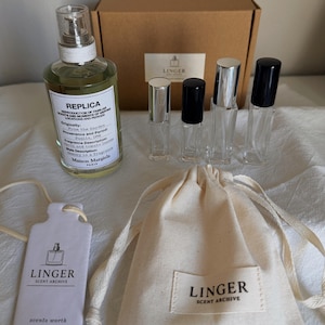May include: A Maison Margiela Replica perfume bottle, four small clear glass spray vials with silver and black caps, a brown cardboard box, a white rectangular tag, and a cream-colored drawstring pouch, all featuring Linger Scent Archive branding.