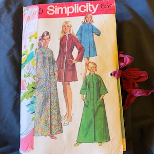 May include: A Simplicity sewing pattern envelope featuring illustrations of four different robe styles in floral, maroon, blue, and green colors. The envelope has a pink fabric strip attached to the left side and a dark pink ribbon tied on the right side.