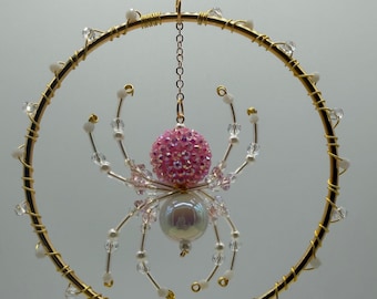 May include: A hanging ornament featuring a gold wire hoop decorated with small clear beads, suspending a spider crafted from wire, a pink glittery spherical bead for the body, and a smooth iridescent white bead for the abdomen.