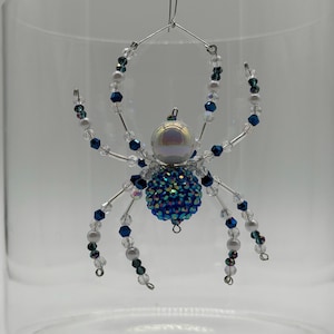 May include: A decorative spider ornament crafted from wire and beads. The body features a large iridescent white sphere and a smaller sphere covered in blue and green faceted crystals. Eight legs are adorned with clear, blue, and white beads.