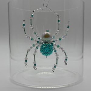 May include: A decorative spider ornament crafted from wire and beads. The spider features a large, iridescent white pearl-like head and a round, textured body made of small, bright turquoise beads. Eight legs are constructed with clear and turquoise beads.