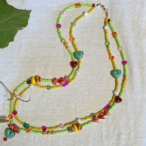 May include: A multi-strand beaded necklace featuring a vibrant mix of lime green and yellow seed beads. The design incorporates various accent beads, including light blue heart-shaped charms, red flower-shaped beads, and small white marble-like spheres.