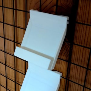 Könnte beinhalten: Zwei weiße, 3D-gedruckte Wandregale. Die rechteckigen Regale haben eine Lippe an der Vorderseite und sind an einem schwarzen Drahtgitter vor einem Holzgrund befestigt. Die Regale sind für kleine Gegenstände konzipiert.