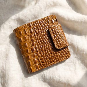Op de afbeelding: Een vierkante bruine portemonnee met een opvallende reliëftextuur van krokodillenleer. De portemonnee is voorzien van een bijpassende klepsluiting aan de rechterkant, met zichtbare stiksels langs de randen en een glanzende afwerking over het gehele oppervlak.