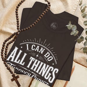 Könnte beinhalten: Ein dunkelbraunes T-Shirt mit weißem grafischem Text, der I CAN DO ALL THINGS THROUGH CHRIST lautet, begleitet von einem Rosenkranz aus Holzperlen mit Kreuzanhänger. Das T-Shirt hat ein kreisförmiges Logo am inneren Halsausschnitt Still & Faith Co.