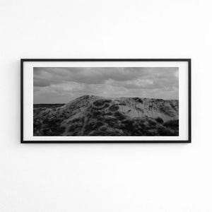 Può includere: Una fotografia di paesaggio in bianco e nero all'interno di una sottile cornice nera. L'immagine raffigura una duna di sabbia coperta da ciuffi d'erba sotto un cielo nuvoloso. La composizione è orizzontale, catturando la trama del terreno.