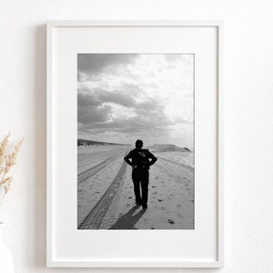 Può includere: Fotografia in bianco e nero incorniciata di una persona su una spiaggia. La persona, vestita di scuro, è in piedi con le mani sui fianchi. Le tracce di pneumatici segnano la sabbia, che conduce verso l'orizzonte sotto un cielo nuvoloso.