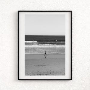 Può includere: Una stampa artistica da parete incorniciata in bianco e nero che raffigura una figura solitaria che cammina lungo una spiaggia sabbiosa. La composizione cattura le onde dell'oceano sullo sfondo sotto un cielo limpido, racchiusa in una cornice nera.