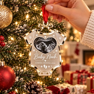 May include: A gold-rimmed, babygrow-shaped Christmas ornament featuring a central heart-shaped ultrasound image. The ornament displays the text 'Baby Noah' and 'COMING WINTER 2026' with snowflake and footprint icons, suspended by a red ribbon.