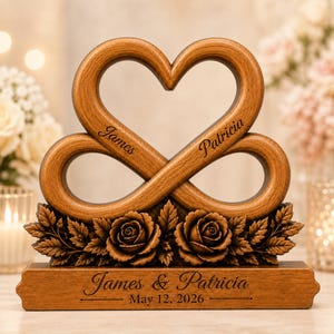 May include: A wooden heart-shaped sculpture featuring an infinity loop design with the names James and Patricia engraved on the upper curves. The base is adorned with carved roses and leaves, and includes the text James & Patricia May 12, 2026.