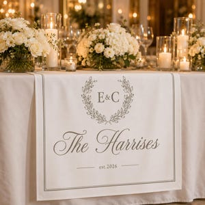 May include: A white fabric table runner featuring a gray printed design with a laurel wreath surrounding the letters E&C, the text The Harrises, and the date est.2026. The runner is draped over a table with white floral centerpieces and candles.
