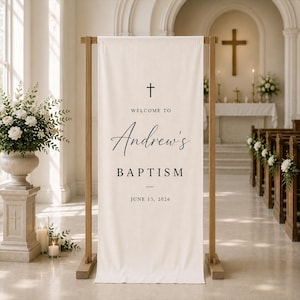 Pode incluir: Uma faixa alta de tecido branco sujo suspensa numa moldura de madeira clara. A faixa apresenta um ícone de cruz preta, o texto "WELCOME TO", o nome "Andrew's" em letra cursiva, "BAPTISM" em letras de forma e a data "JUNE 13, 2026".