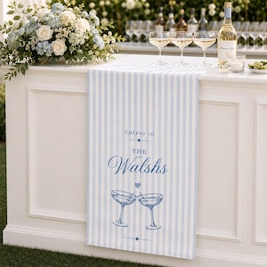 Könnte beinhalten: Ein hellblau-weiß gestreifter Tischläufer mit dem blauen Schriftzug "CHEERS TO THE Walshs", ergänzt durch eine Grafik von zwei anstoßenden Cocktailgläsern und einem kleinen Herzsymbol, drapiert über einer weißen Bar mit Blumendekoration.