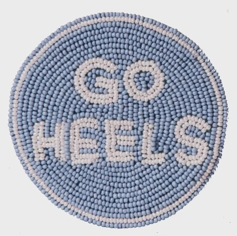 May include: Round, beaded coaster in light blue with the words "GO HEELS" in white beads. The coaster has a white beaded border. This decorative item is perfect for any fan.