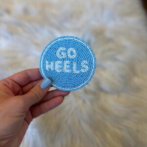 May include: A round, light blue beaded coaster with the words "GO HEELS" in white beads. The coaster has a white beaded border and is held up by a hand with light blue nail polish.