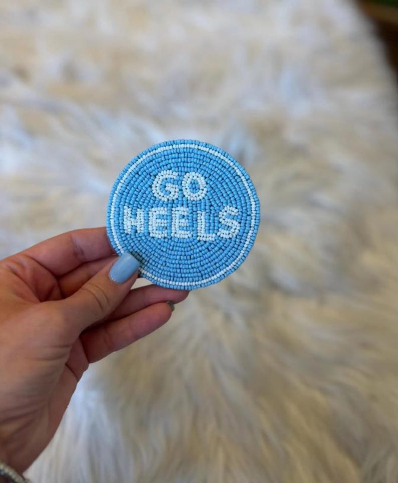 May include: A round, light blue beaded coaster with the words "GO HEELS" in white beads. The coaster has a white beaded border and is held up by a hand with light blue nail polish.