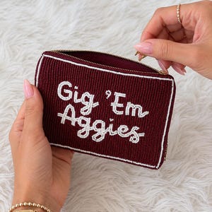 May include: A rectangular maroon beaded pouch featuring the white text Gig 'Em Aggies. The pouch has a gold zipper closure along the top edge and is accented with thin white beaded border lines along the top and bottom edges of the front panel.
