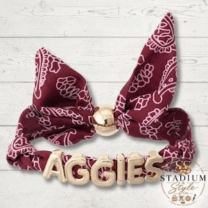 May include: A maroon fabric hair scrunchie featuring a white paisley pattern and a large bow at the top. The scrunchie is accented with a central gold-toned bead and gold-toned letter beads that spell out the word AGGIES in a rounded font.