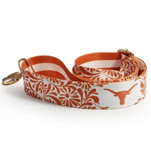 May include: A white and burnt orange patterned bag strap featuring a repeating floral design and a longhorn steer logo. The strap is constructed from woven fabric and includes two gold-toned metal swivel lobster claw clasps at each end.