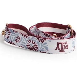 Op de afbeelding: Een witte stoffen riem met een bordeauxrood en lichtgrijs bloemenpatroon met het bordeauxrode ATM-logo van de Texas A&M University. De riem is voorzien van goudkleurige metalen hardware, waaronder verstelbare schuifgespen en draaibare clips.