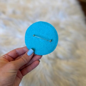 May include: A round, bright turquoise felt pin with a silver safety pin clasp on the back. The pin is a solid color and appears to be handmade. The background is a soft, white, textured surface.