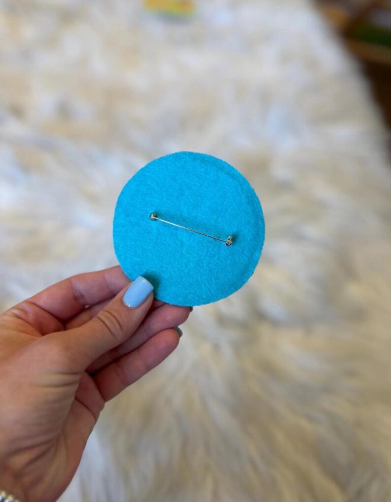 May include: A round, bright turquoise felt pin with a silver safety pin clasp on the back. The pin is a solid color and appears to be handmade. The background is a soft, white, textured surface.