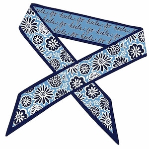 Op de afbeelding: Een lange, smalle sjaal met een lichtblauwe achtergrond en een herhalend donkerblauw en wit bloemenpatroon. E&eacute;n kant toont de herhaalde cursieve tekst "go heels" in het donkerblauw, terwijl de andere kant een dicht bloemenmotief heeft.