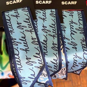 Op de afbeelding: Drie lichtblauwe smalle sjaals met donkerblauwe randen, elk bevestigd aan een zwarte kopkaart met de tekst SKINNY SCARF. De stof heeft een herhalend donkerblauw cursief schrift met de tekst go heels en een bloemenpatroon aan de onderrand.