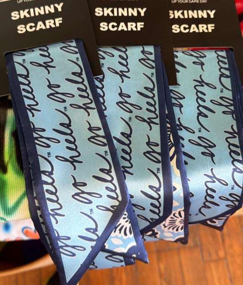 Op de afbeelding: Drie lichtblauwe smalle sjaals met donkerblauwe randen, elk bevestigd aan een zwarte kopkaart met de tekst SKINNY SCARF. De stof heeft een herhalend donkerblauw cursief schrift met de tekst go heels en een bloemenpatroon aan de onderrand.