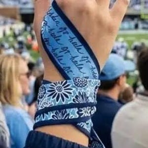 Op de afbeelding: Een blauw-witte stoffen polswikkel met een herhalend bloemmotief en een gedeelte met cursieve tekst. De stof is om de pols en handpalm gewikkeld en vastgezet met een knoop, waarbij een combinatie van licht- en donkerblauwe tinten zichtbaar is.
