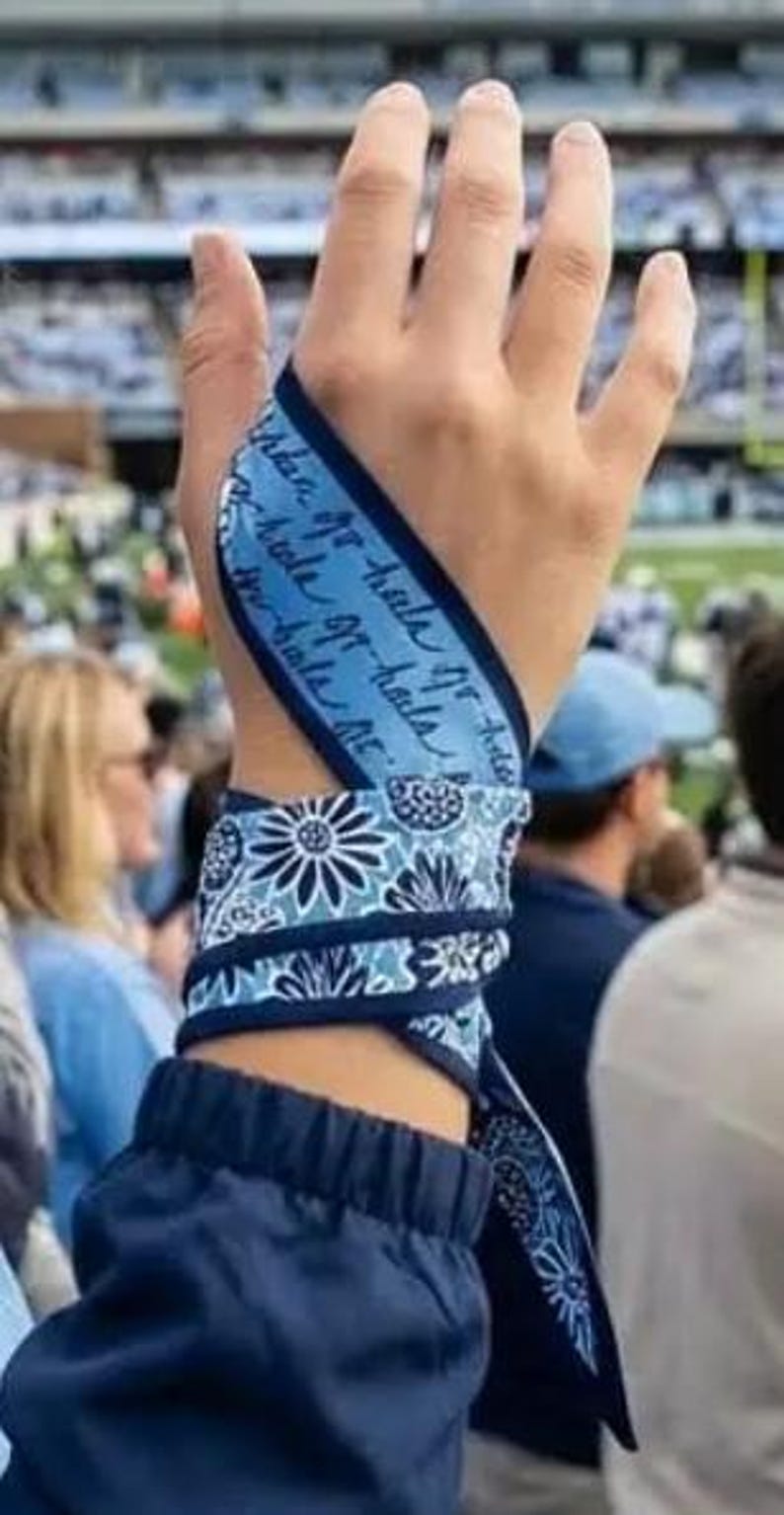 Op de afbeelding: Een blauw-witte stoffen polswikkel met een herhalend bloemmotief en een gedeelte met cursieve tekst. De stof is om de pols en handpalm gewikkeld en vastgezet met een knoop, waarbij een combinatie van licht- en donkerblauwe tinten zichtbaar is.