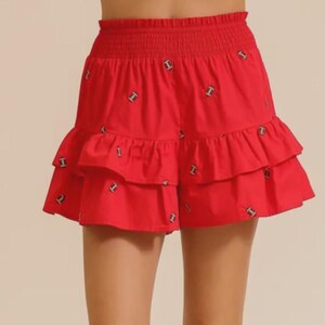 Puede incluir: Una falda de color rojo brillante con cintura elástica fruncida y dos capas de volantes. La tela está decorada con un patrón repetitivo de pequeños iconos de balones de fútbol americanos en color marrón y blanco sobre la superficie.
