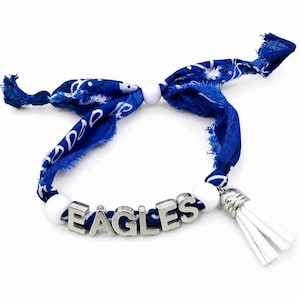 Op de afbeelding: Een blauwe stoffen armband met een wit paisley-patroon, vastgeknoopt. De armband bevat zilverkleurige metalen letterkralen die EAGLES spellen, twee witte ronde kralen en een witte kunstlederen kwast bevestigd met een zilveren dop.